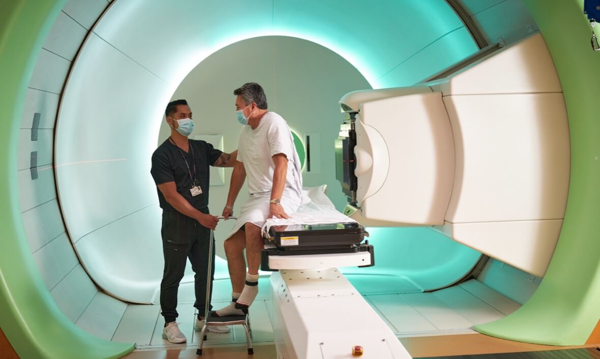 Medical worker assists seated patient inside circular machine that resembles a large MRI enclosure.
