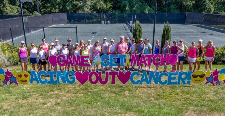A large group stands by tennis courts with signs reading Game, Set, Match and Acing Out Cancer.